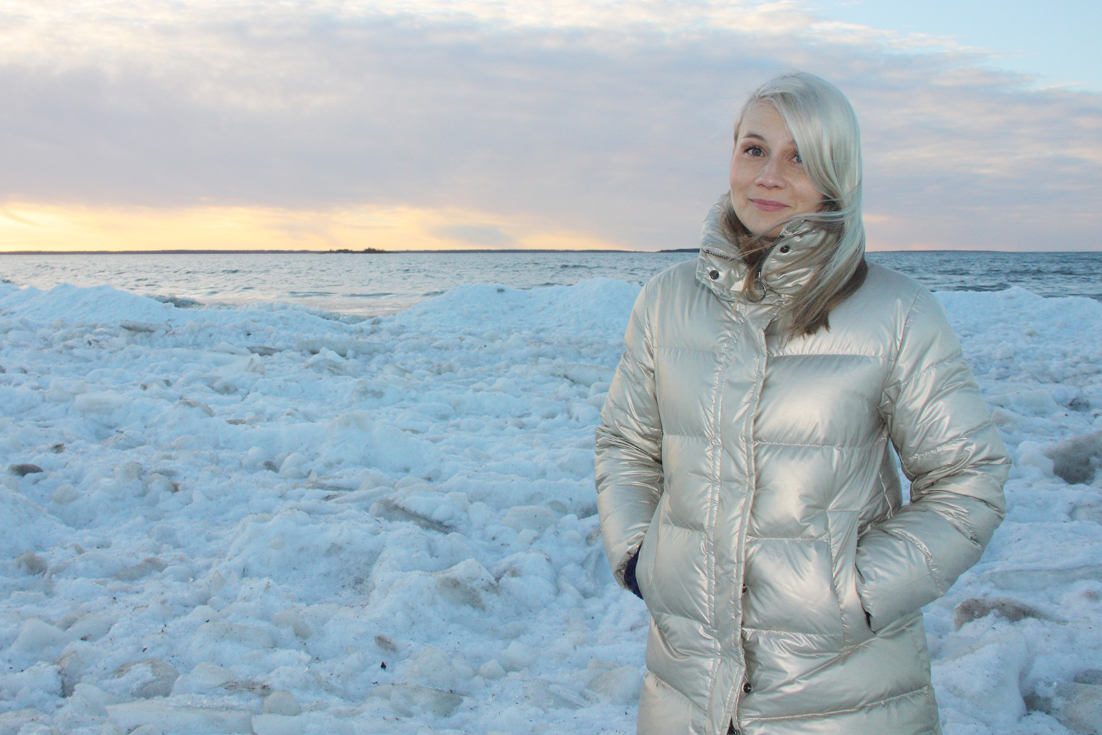 Vaaleahiuksinen Jenny Simojoki on pukeutunut kullanväriseen untuvatakkiin ja katsoo kameraan kädet taskussa. Takana on talvista maisemaa, meri ja auringonlaskun värejä.