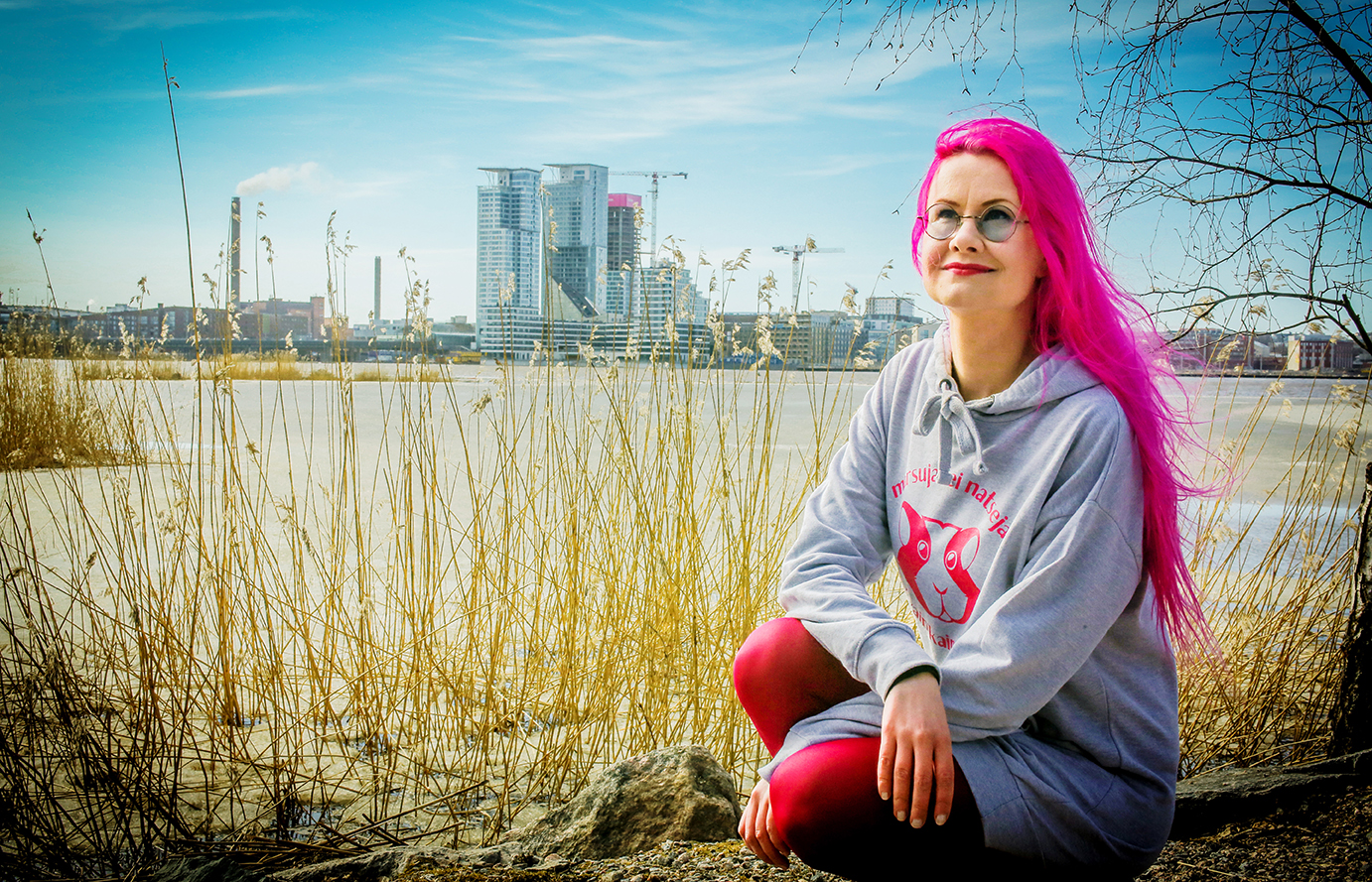 Pinkkihiuksinen henkilö istuu kyykyssä rannalla. Taustalla on heinikkoa ja vastarannalla näkyy kerrostaloja.