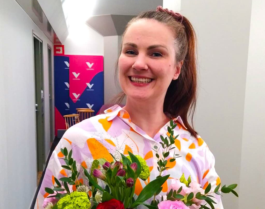 Iloinen pitkähiuksinen henkilö katsoo kameraan kukkakimppu sylissään.