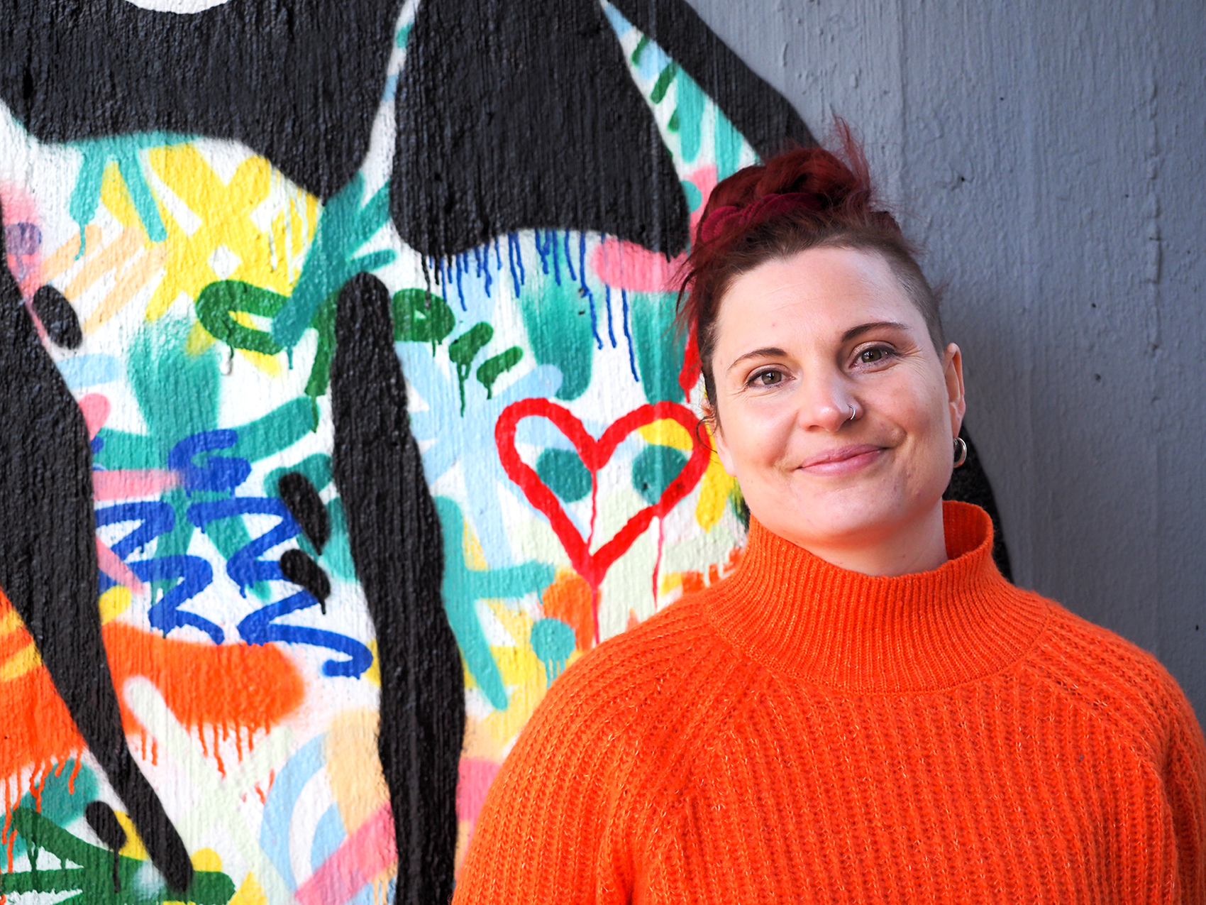 Oranssiin neuleeseen pukeutunut Annuska Dal Maso seisoo värikkään maalatun seinän edessä ja katsoo hymyillen kameraan.