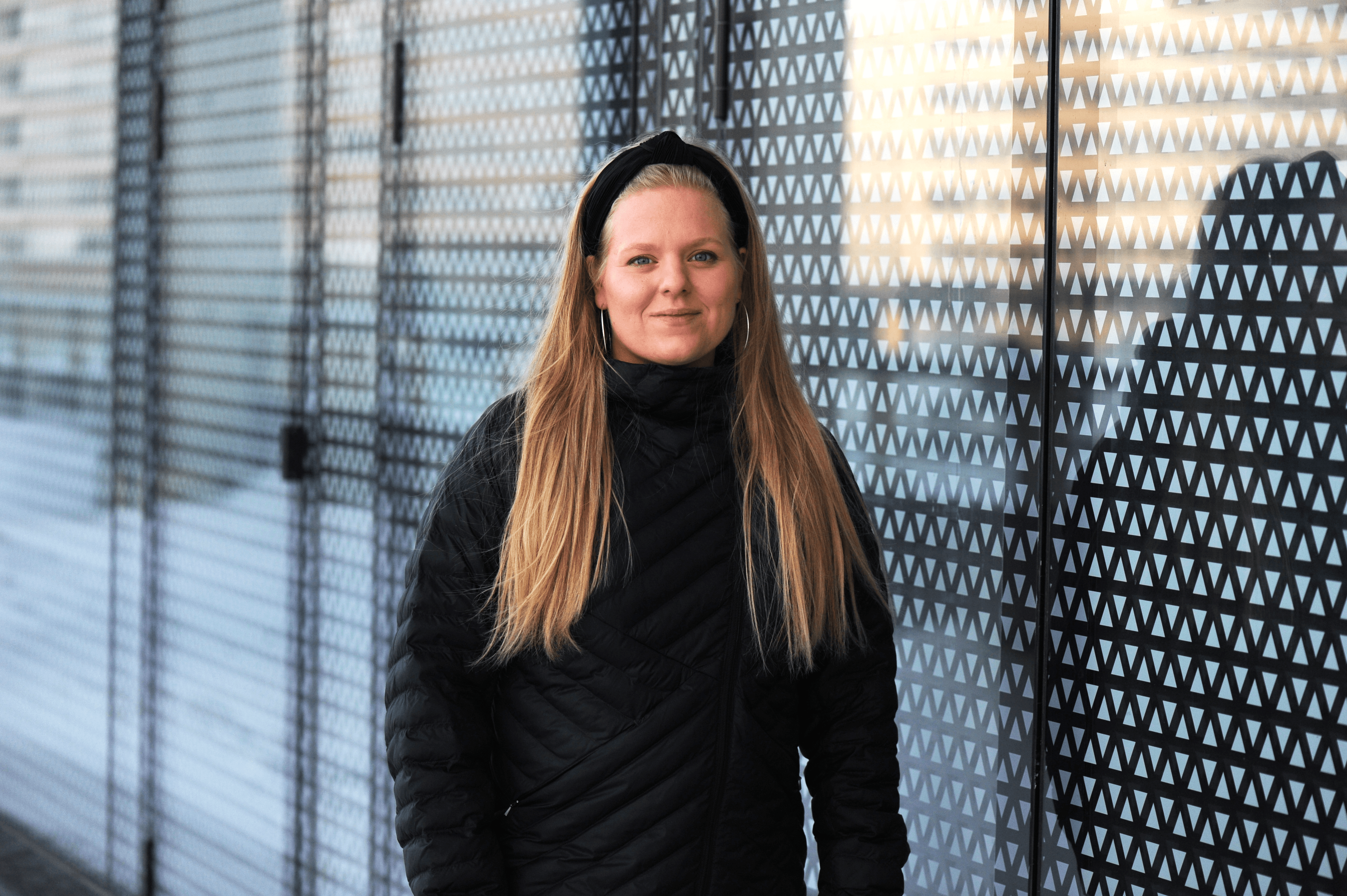 Mustiin pukeutunut vaaleahiuksinen henkilö seisoo lasiseinän edessä ja katsoo kameraan.