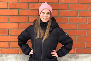 Elina Rodriguez on pukeutunut mustaan toppatakkiin ja vaaleanpunaiseen pipoon. Hän seisoo oranssin tiiliseinän edessä kädet vyötäröllä.