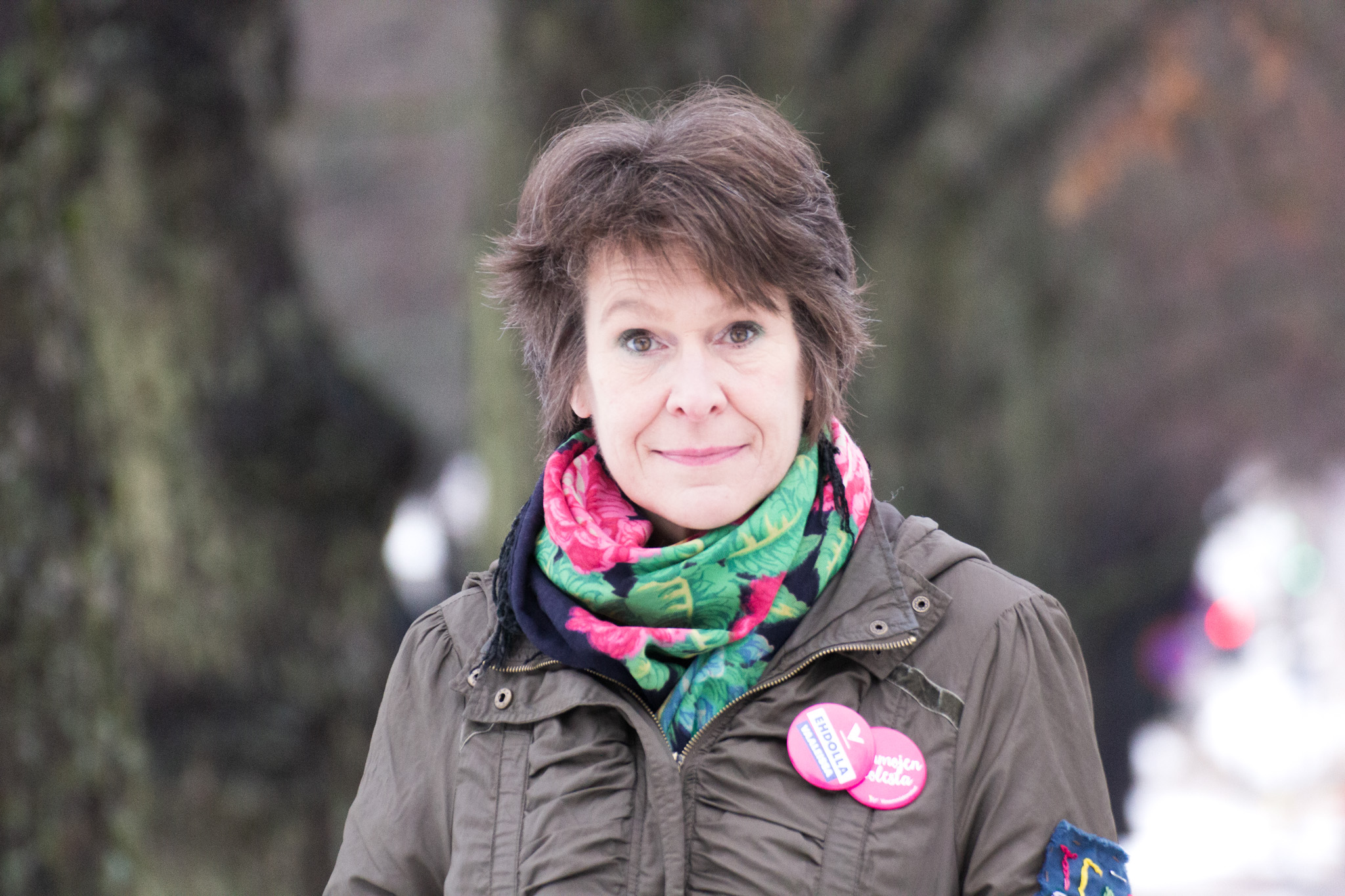 Arja-Tauria Huttunen on valkoihoinen ja hänellä on lyhyet ruskeat hiukset. Hänellä on vihreä takki, jossa on kaksi pinkkiä rintamerkkiä, kaulassa kukallinen huivi. Hän katsoo vakavana suoraan kameraan. Taustalla näkyy epätakkana puita.