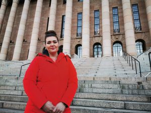 Satu Viksten seisoo eduskuntatalon portaillla. Hän on valkoinen, tummahiuksinen ja pukeutunut kirkkaanpunaiseen takkiin.