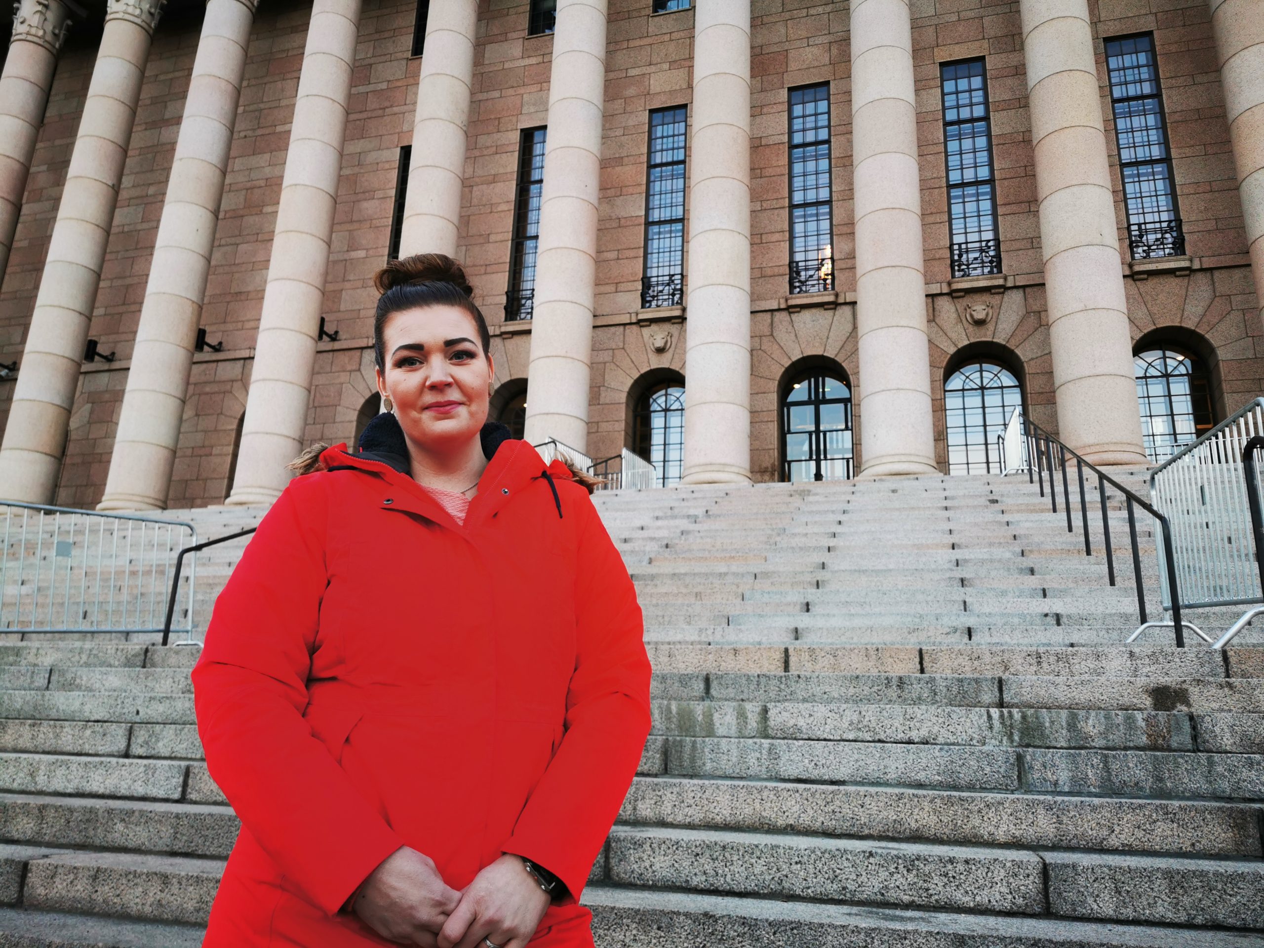 Satu Viksten seisoo eduskuntatalon portaillla. Hän on valkoinen, tummahiuksinen ja pukeutunut kirkkaanpunaiseen takkiin.