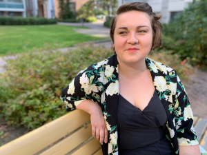 Mustakukalliseen asuun pukeutunut henkilö istuu puistonpenkillä ja katsoo sivuttain kameran ohi. Taustalla näkyy nurmikkoa, pensaita, kävelytie ja taloja.