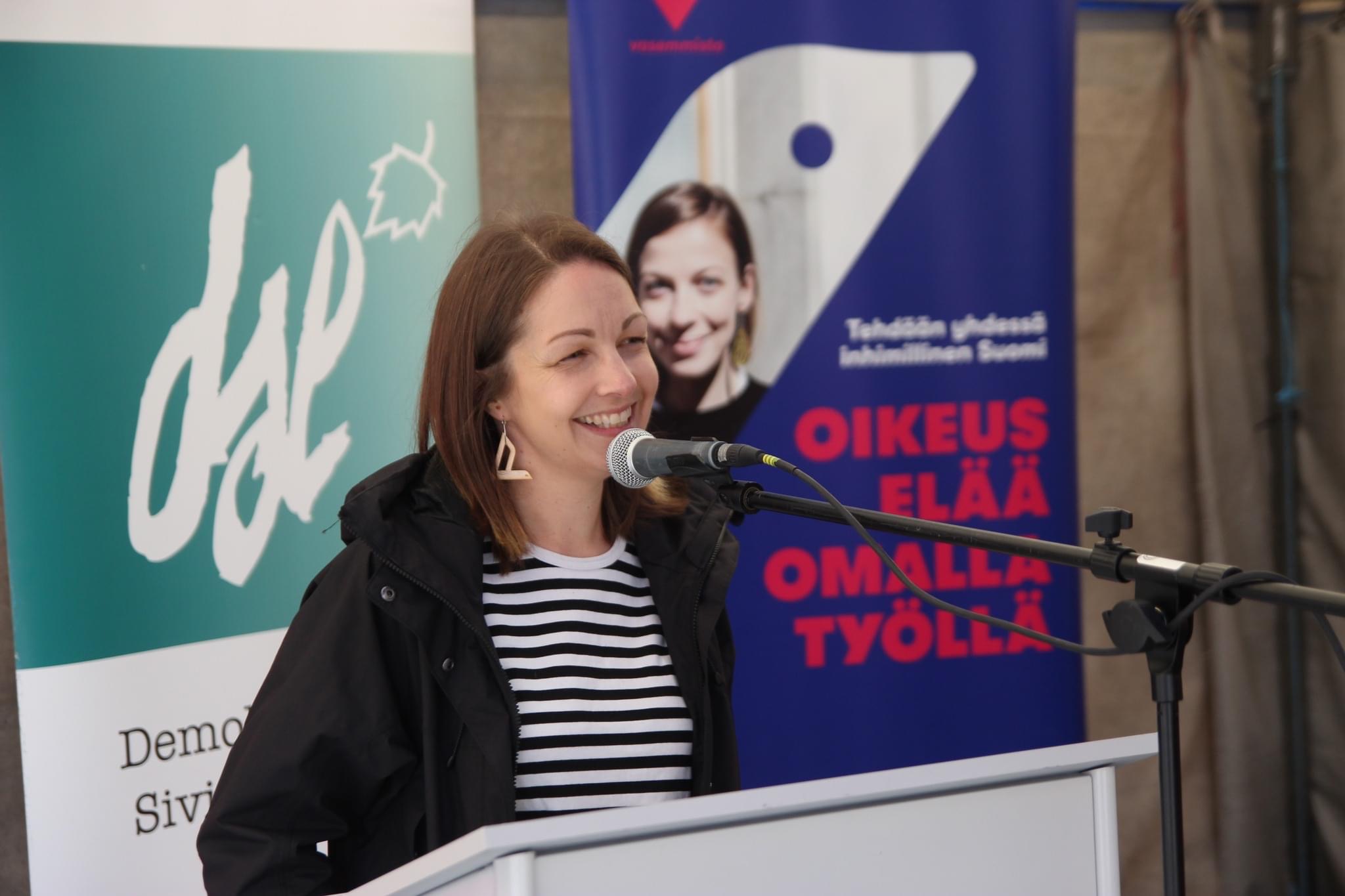 Piia Kilpeläinen-Tuoma seisoo puhujankorokkeen takana ja puhuu hymyillen mikrofoniin. Hän on valkoinen ja ruskeahiuksinen ja hänellä on yllään musta takki ja mustavalkoraidallinen paita. Taustalla on kaksi roll-upia.