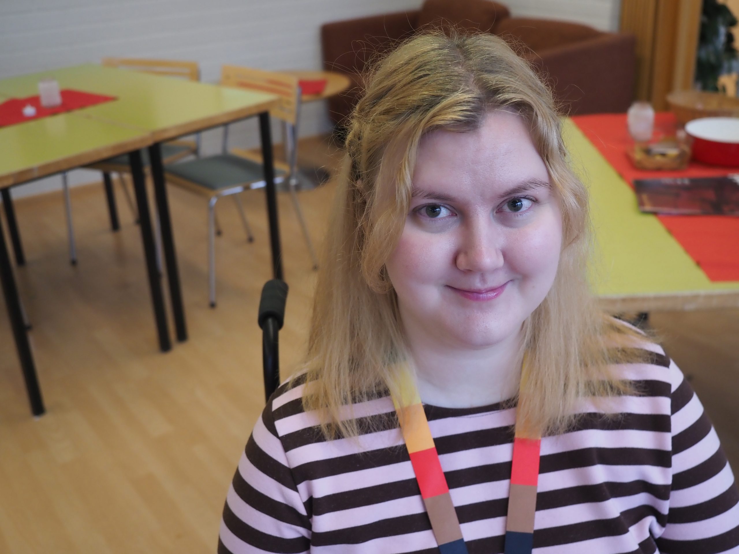 Lähikuva Katariina Aakusista, joka katsoo hymyillen kameraan. Aakusilla on raidallinen paita ja värikäs kaulanauha. Taustalla on kaksi pöytää ja sohva.