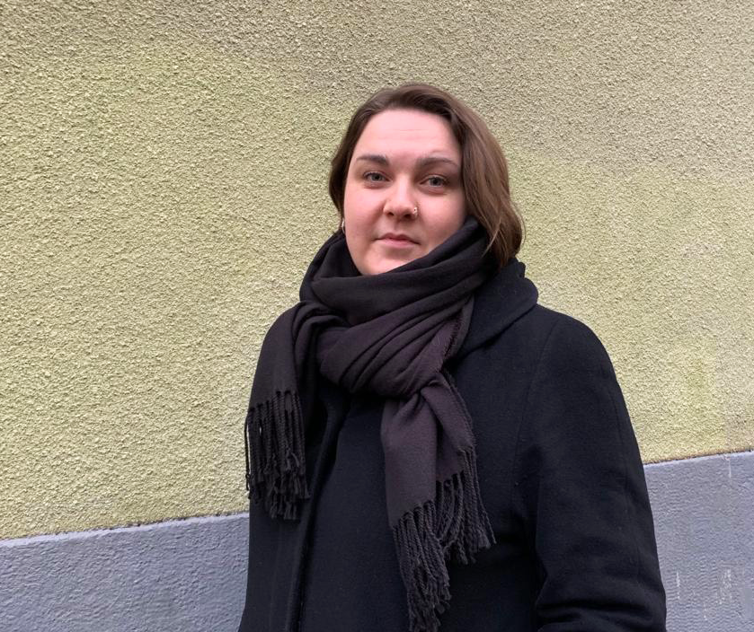 Mustaan takkiin ja kaulahuiviin pukeutunut henkiläö seisoo keltaisen kiviseinän edessä.