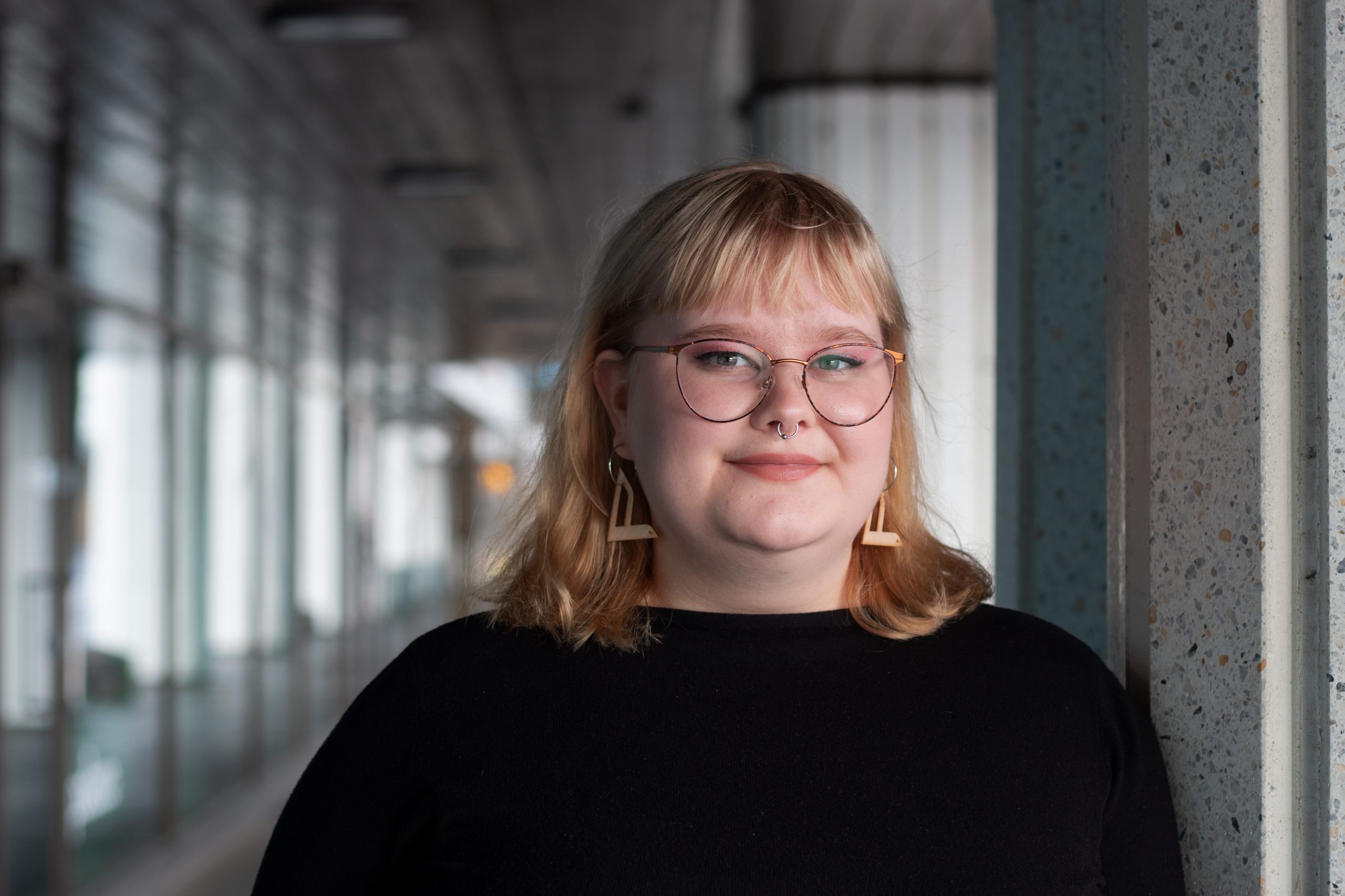 Valkoinen ja vaaleahiuksinen Selmi Holopainen katsoo suoraan kameraan. Hänellä on silmälasit, musta paita ja vasemmistoliiton lintukorvikset. Taustalla näkyy käytävää ja ikkunoita.