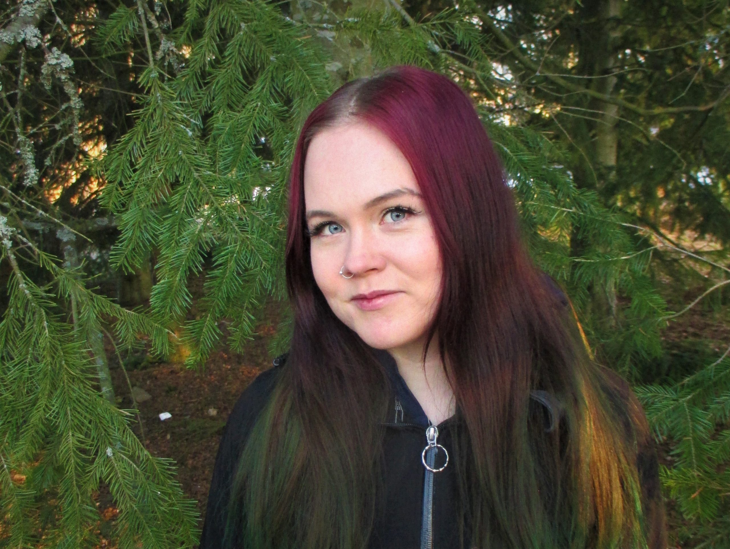 Noora Kyllönen hymyilee ja katsoo kameraan hieman sivuittain. Taustalla on vihreä havupuu.