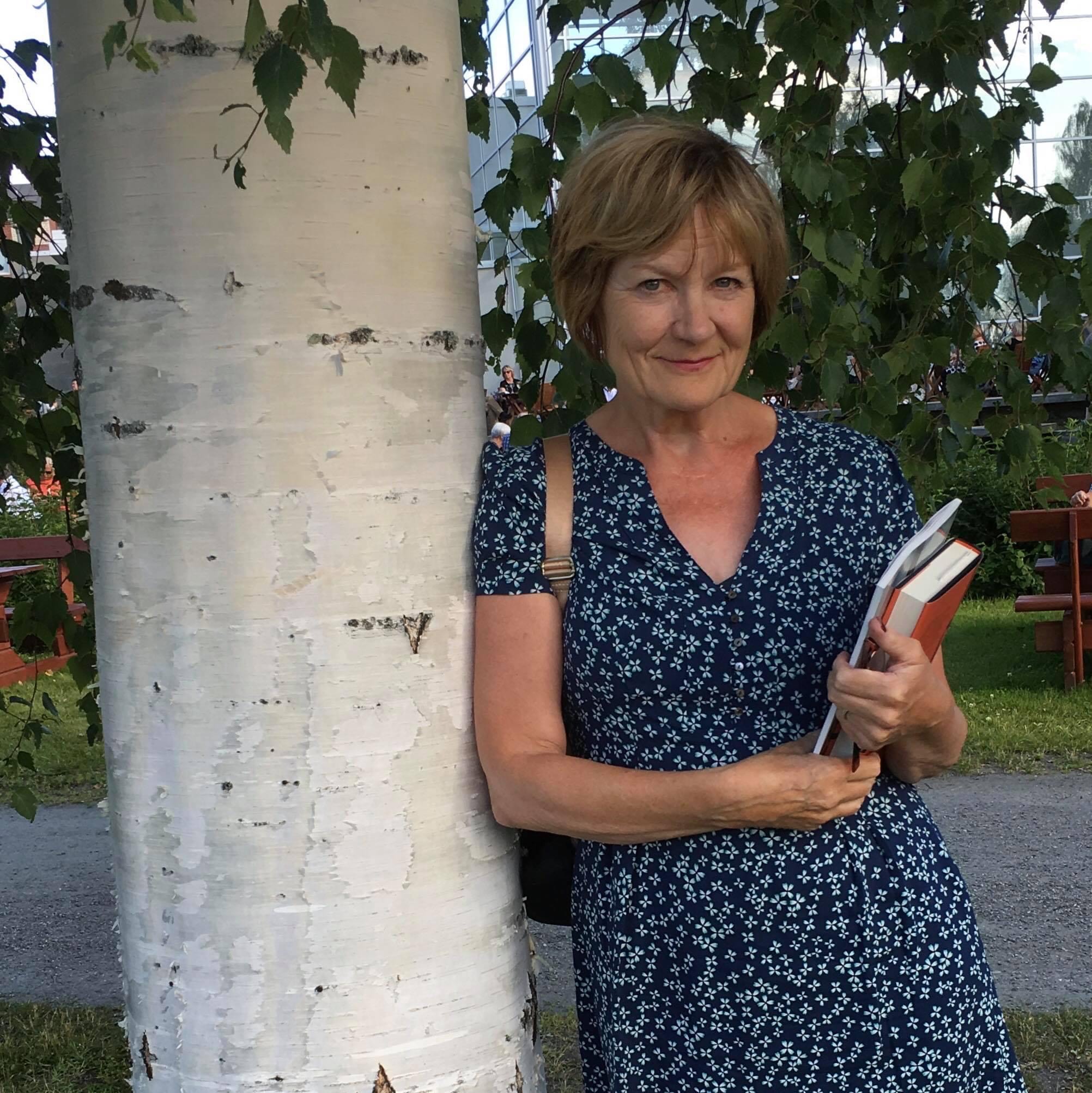 Ulla-Leena Alppi katsoo hymyillen kameraan, nojaa koivunrunkoon ja pitelee kädessään pientä kirjapinoa.