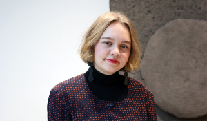 Tummiin vaatteisiin pukeutunut henkilö katsoaa hymyillen kameraan. Taustalla näkyy valkoista ja ruskeaa seinää.