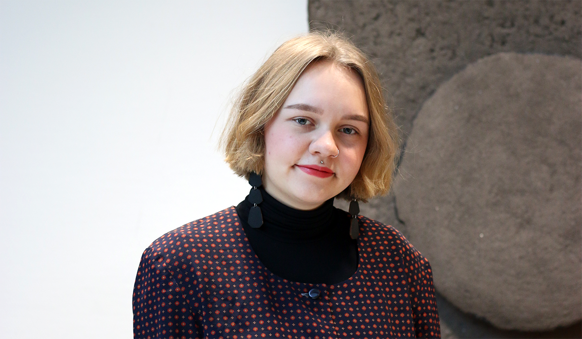 Tummiin vaatteisiin pukeutunut henkilö katsoaa hymyillen kameraan. Taustalla näkyy valkoista ja ruskeaa seinää.