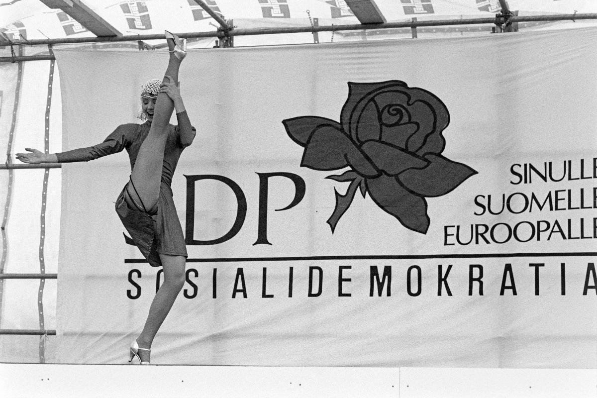 Mustavalkoinen valokuva, jossa etualalla jalkaa nostava can-can-tanssija ja taustalla banderolli, jossa teksti: "SDP Sinulle Suomelle Euroopalle Sosialidemokratia"