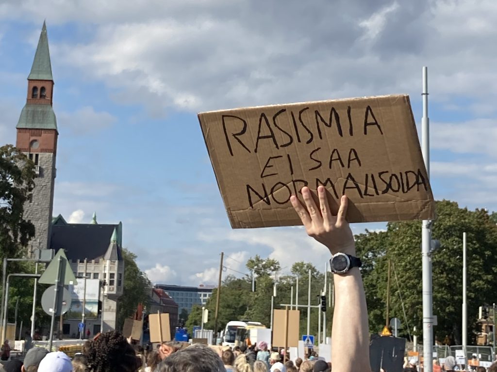 Mielenosoituskyltti taivasta vasten. Teksti "Rasismia ei saa normalisoida.