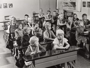 Mustavalkoinen valokuva, jossa lapsia istumassa pulpettien ääressä koululuokassa.