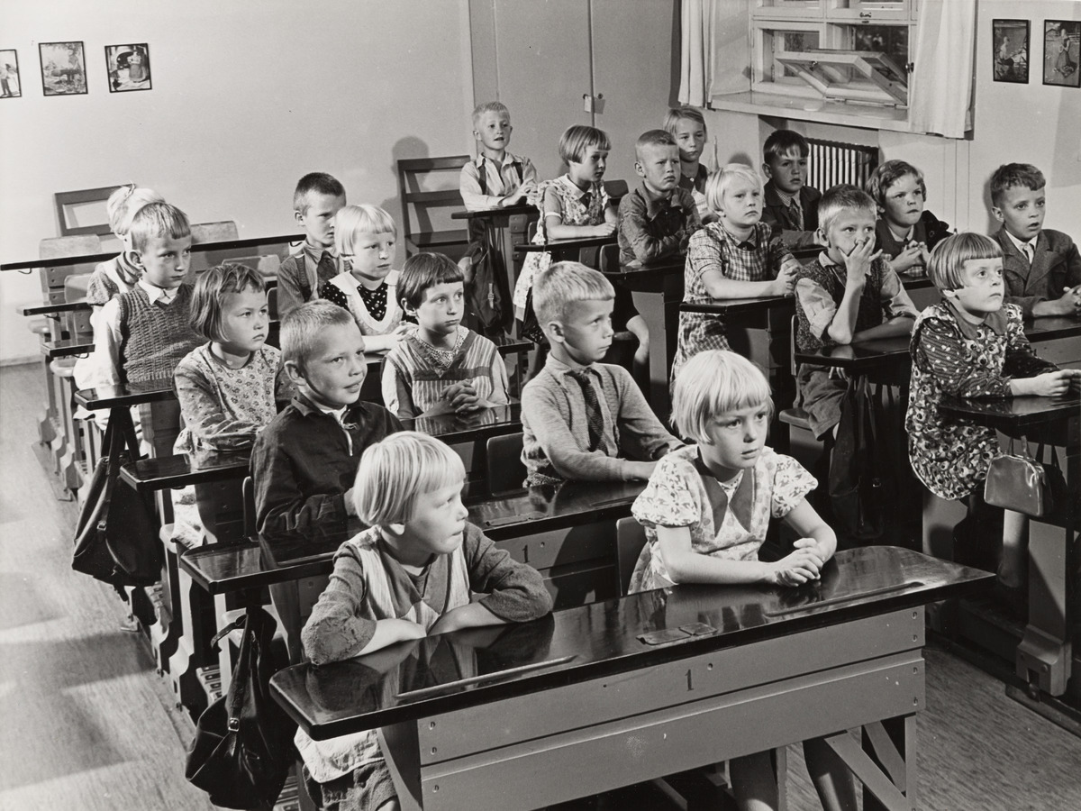 Mustavalkoinen valokuva, jossa lapsia istumassa pulpettien ääressä koululuokassa.
